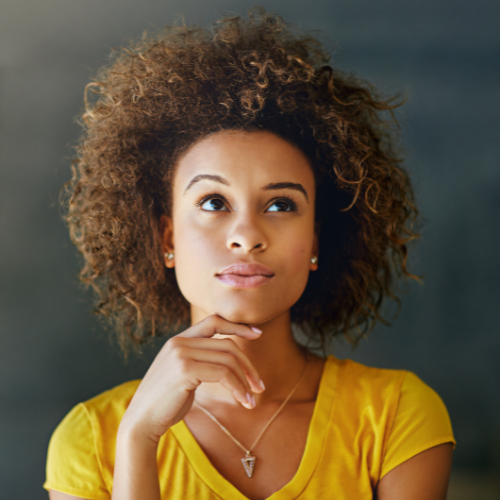 Une femme aux cheveux bouclés, portant une chemise jaune et un collier, regarde vers le haut d'un air pensif, le menton posé sur sa main, sur un fond sombre et flou.